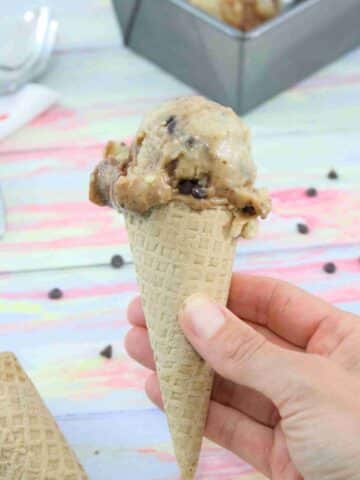 A chocolate swirled protein ice cream in an ice cream cone bring held up close with a baking load of protein ice cream behind it