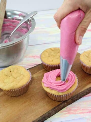 Pink dairy-free cream cheese frosting being piped onto vanilla cupcakes on a cutting board with a steel bowl of frosting behind them.