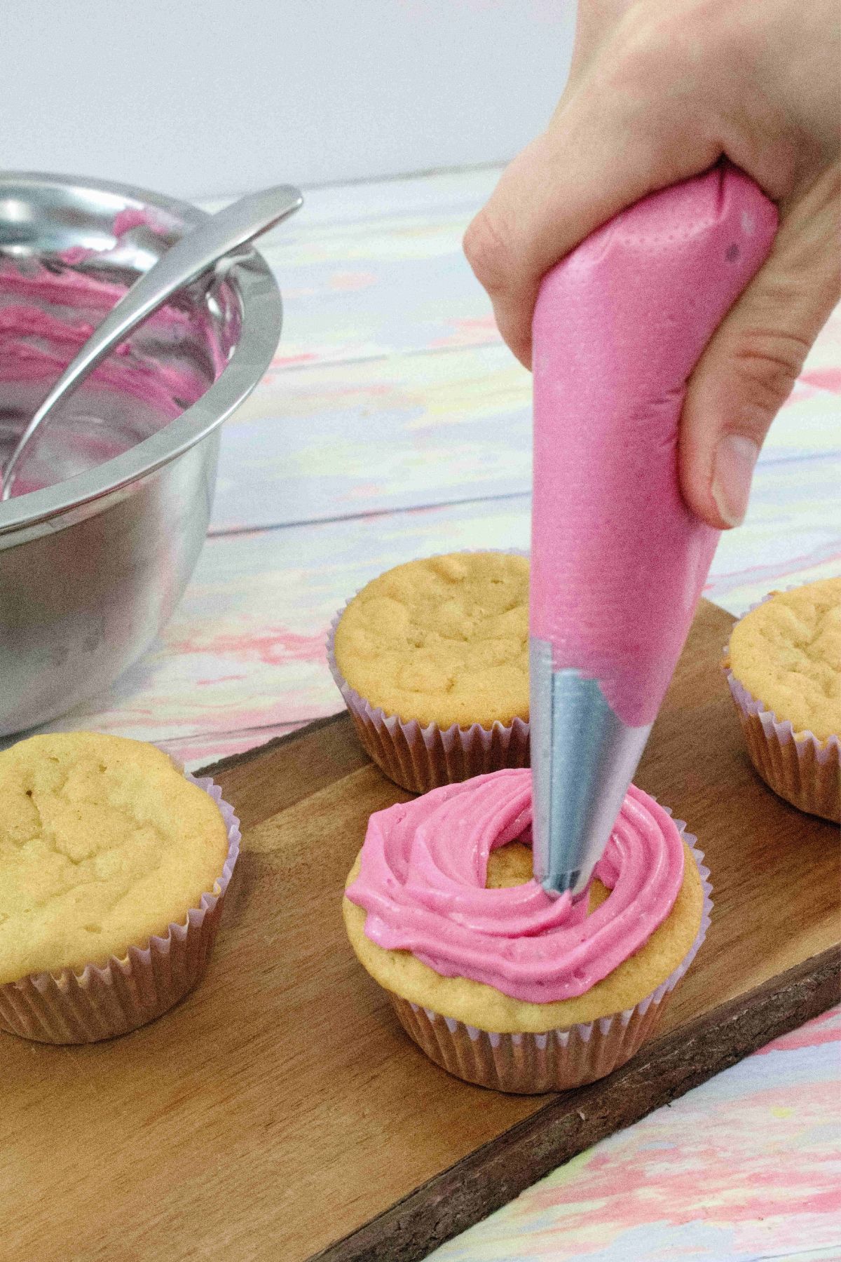 pink dairy-free cream cheese frosting being piped onto vanilla cupcakes on a cutting board with a steel bowl of frosting behind them.