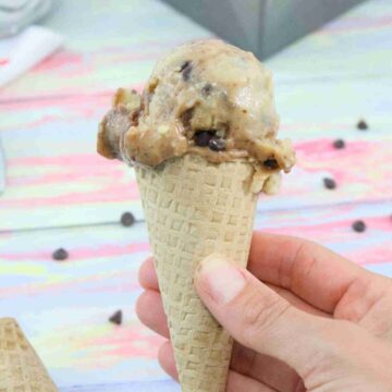A chocolate swirled protein ice cream in an ice cream cone bring held up close with a baking load of protein ice cream behind it.