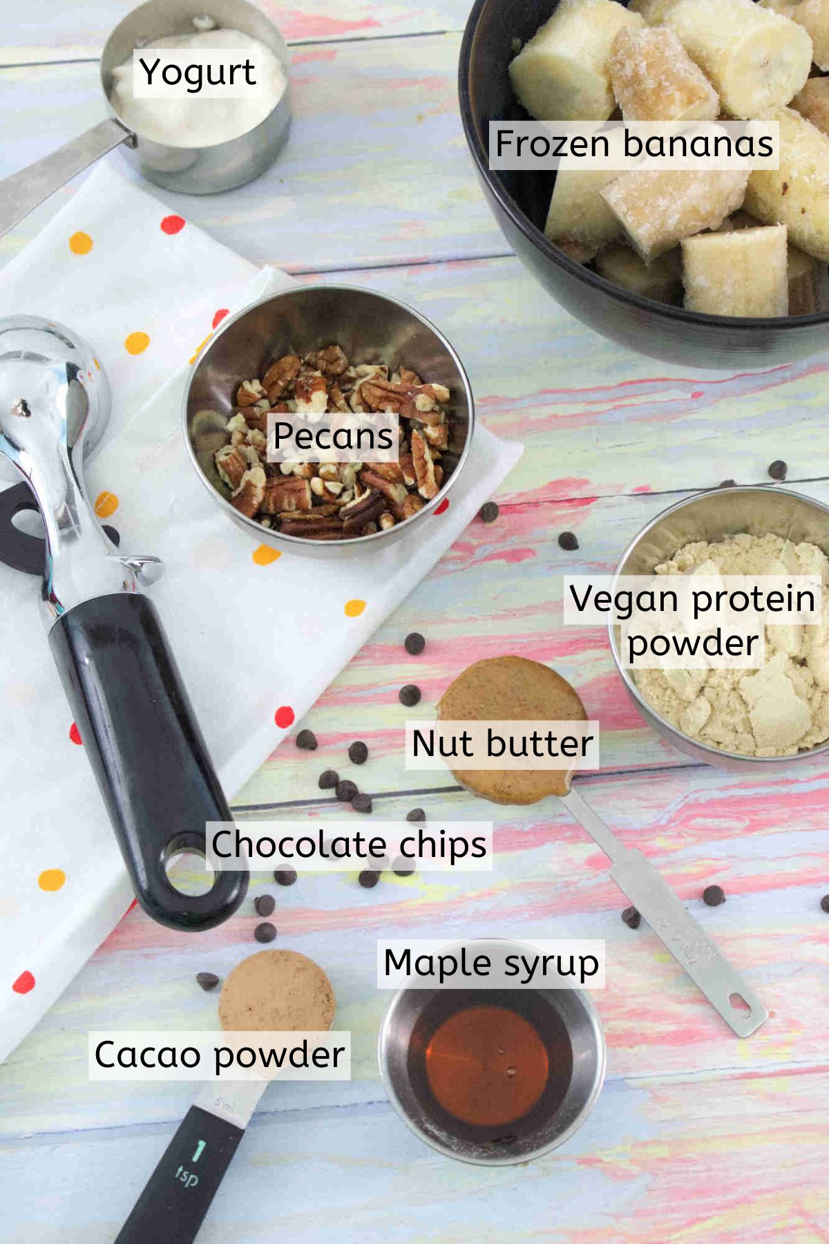 Multiple ingredients to make protein ice cream like frozen bananas, nut butter, chocolate chips and nuts in measuring spoons and cups with text labels over top and an ice cream scoop beside them.