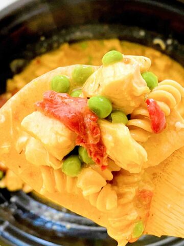 Close up of a wooden spoon with slow cooker chicken alfredo with noodles, peas, chicken and sundried tomatoes on the spoon and the black slow cooker in the background.