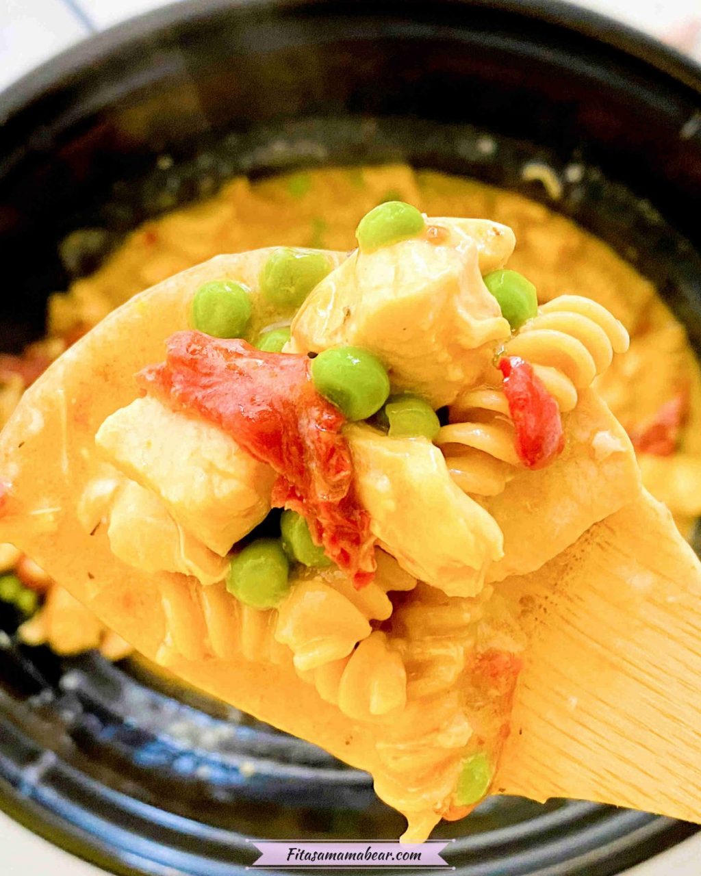 Close up of a wooden spoon with slow cooker chicken alfredo with noodles, peas, chicken and sundried tomatoes on the spoon and the black slow cooker in the background