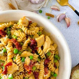 white bowl with chicken pasta with sundried tomatoes and peas and garlic and a light linen around the bowl