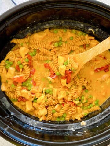 black slow cooker with noodles, sundried tomatoes, chicken, and green peas in a creamy, slightly orange, dairy-free alfredo sauce with a wooden spoon in the slow cooker