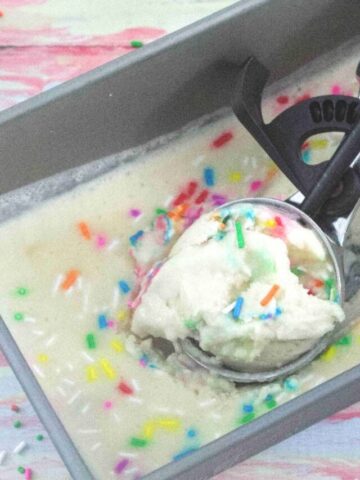 hand using a black and steel ice cream scoop t scoop out vanilla protein ice cream with sprinkles from a baking tin with ice cream cones and sprinkles around it