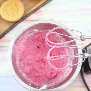 Pink cream cheese frosting in a steel bowl with a handheld blender over top and cupcakes on a cutting board behind them
