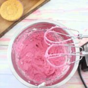 Pink cream cheese frosting in a steel bowl with a handheld blender over top and cupcakes on a cutting board behind them.