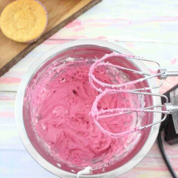 Pink cream cheese frosting in a steel bowl with a handheld blender over top and cupcakes on a cutting board behind them.