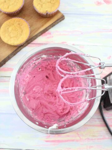 Pink cream cheese frosting in a steel bowl with a handheld blender over top and cupcakes on a cutting board behind them.