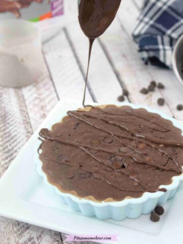 Chocolate protein oatmeal in a blue silicone mold on a white plate being drizzled with melted chocolate with protein powder and chocolate chips behind it