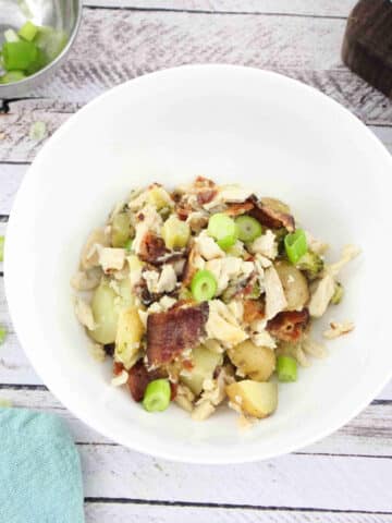 White bowl with chicken, bacon, potato casserole with green onions, blue linen and checkered cutting board behind it