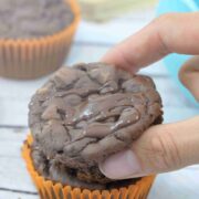 Pin image with text about chocolate protein muffins with two fingers placing a muffin on top of another muffin in an orange liner and more muffins behind it