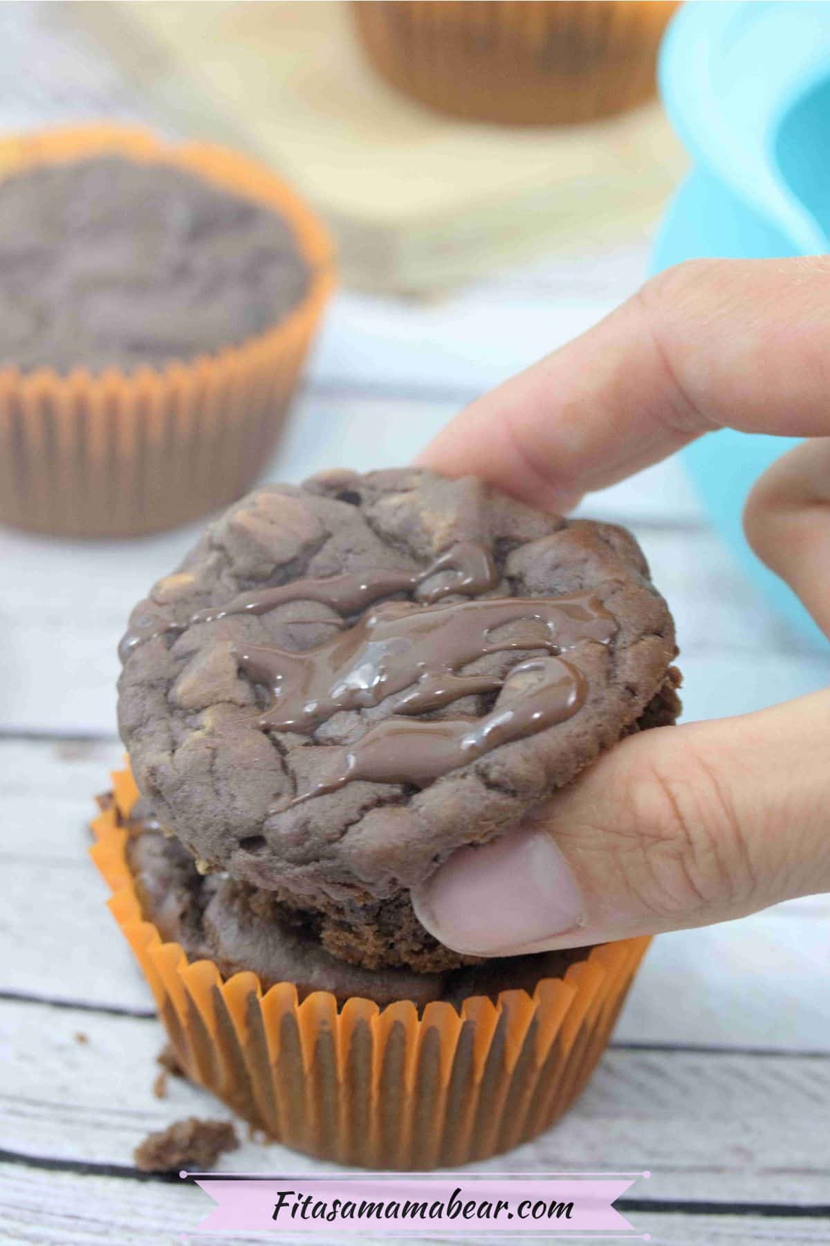 Pin image with text about chocolate protein muffins with two fingers placing a muffin on top of another muffin in an orange liner and more muffins behind it