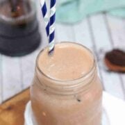 Iced coffee protein shake in a mason jar with blue straws on a cutting board with white cheesecloth and coffee in a french press behind it.