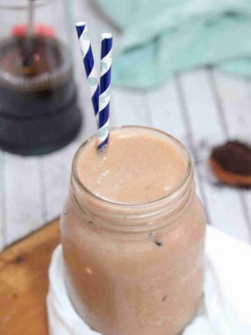 Iced coffee protein shake in a mason jar with blue straws on a cutting board with white cheesecloth and coffee in a french press behind it.