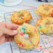 Hand holding a rainbow sprinkled collagen donut over a cooling rack with more donuts behind them.