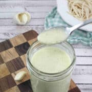 overhead shot of coconut milk alfredo sauce in a mason jar with a spoon coming out of iton a wooden cutting board with garlic, a spoon, and a blue linen around it