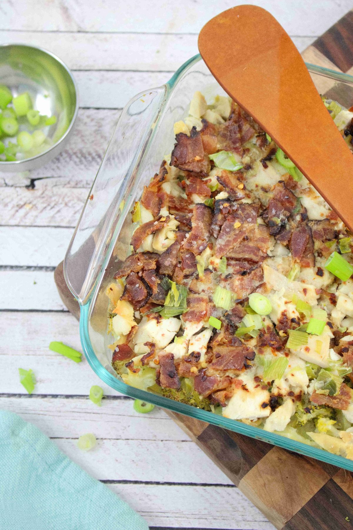 Chicken, bacon and broccoli in a casserole dish on a checkered wooden cutting board with a wooden spoon laying across it and green onions in a bowl and a blue linen beside it.
