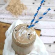 Mason jar with coffee protein shake in it and blue straws on top of white cheesecloth on a cutting board with protein powder and chocolate chips around it.