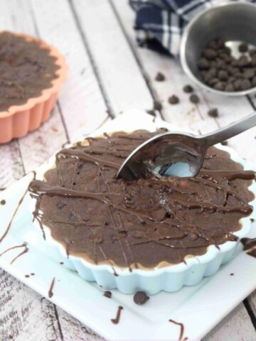 Blue silicone mold with chocolate protein oatmeal with chocolate sauce drizzled on top and a spoon in the oatmeal, chocolat chips in a bowl and another orange mold with oatmeal behind it