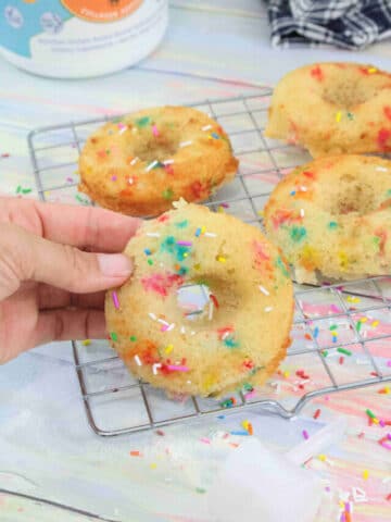 Hand holding a rainbow sprinkled collagen donut over a cooling rack with more donuts behind them.