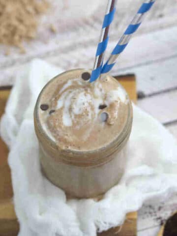 Mason jar with coffee protein shake in it and blue straws on top of white cheesecloth on a cutting board with protein powder and chocolate chips around it.