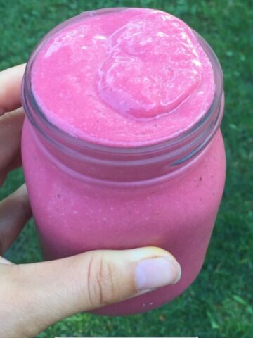 Bright pink smoothie in a mason jar with a hand holding it above grass