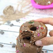 Two fingers placing a chocolate protein donut with sprinkles on top of two more donuts with more behind them.
