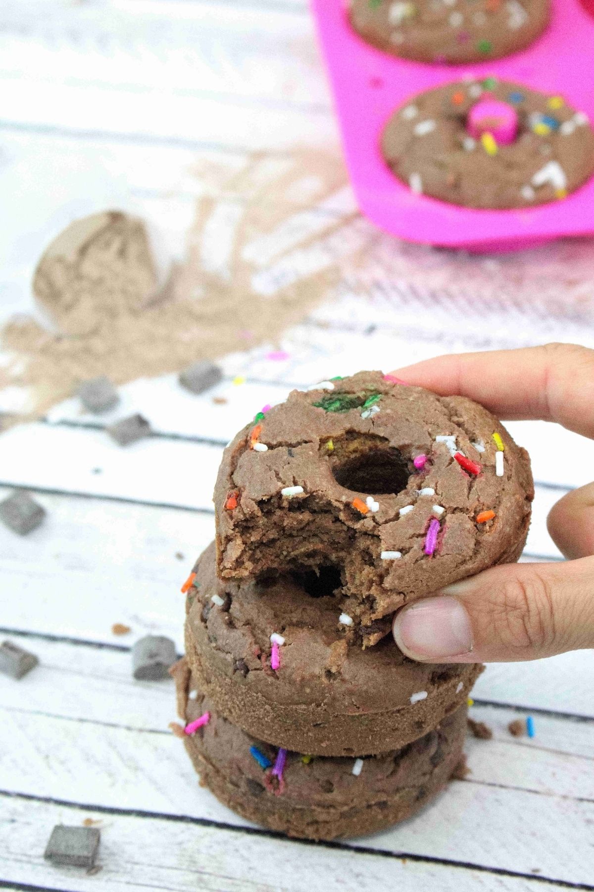 Two fingers placing a chocolate protein donut with sprinkles on top of two more donuts with more behind them.