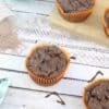 Chocolate muffins in orange liners with some on a wooden cutting board and chocolate sauce drizzled around them with a blue pot of sauce, scoop of protein powder, and a blue linen behind them.