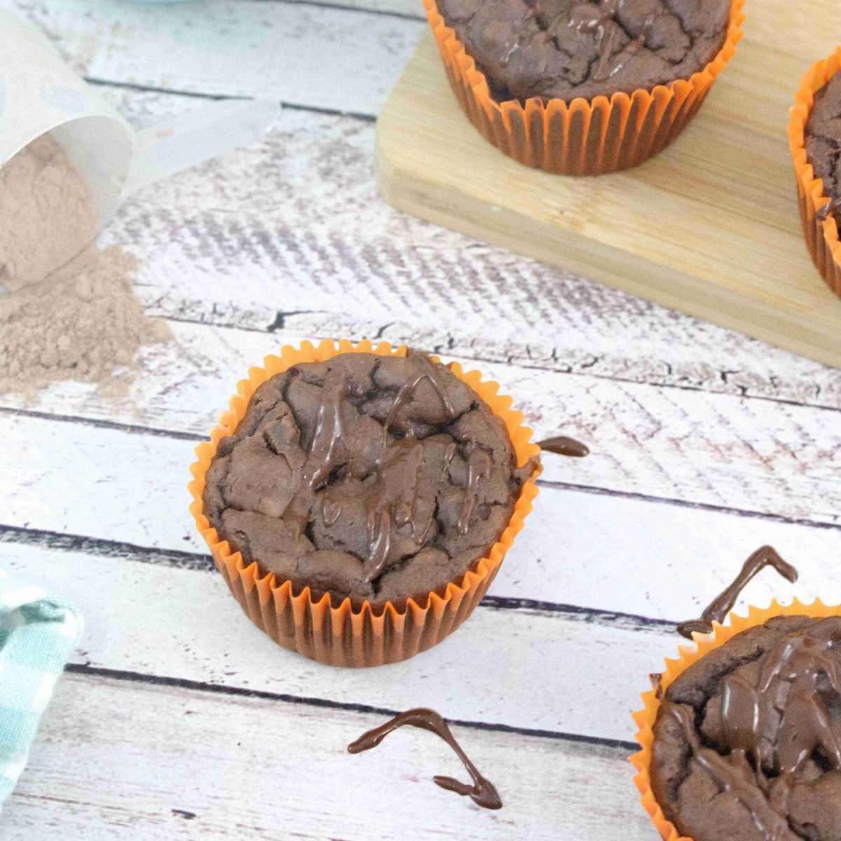 Chocolate muffins in orange liners with some on a wooden cutting board and chocolate sauce drizzled around them with a blue pot of sauce, scoop of protein powder, and a blue linen behind them.