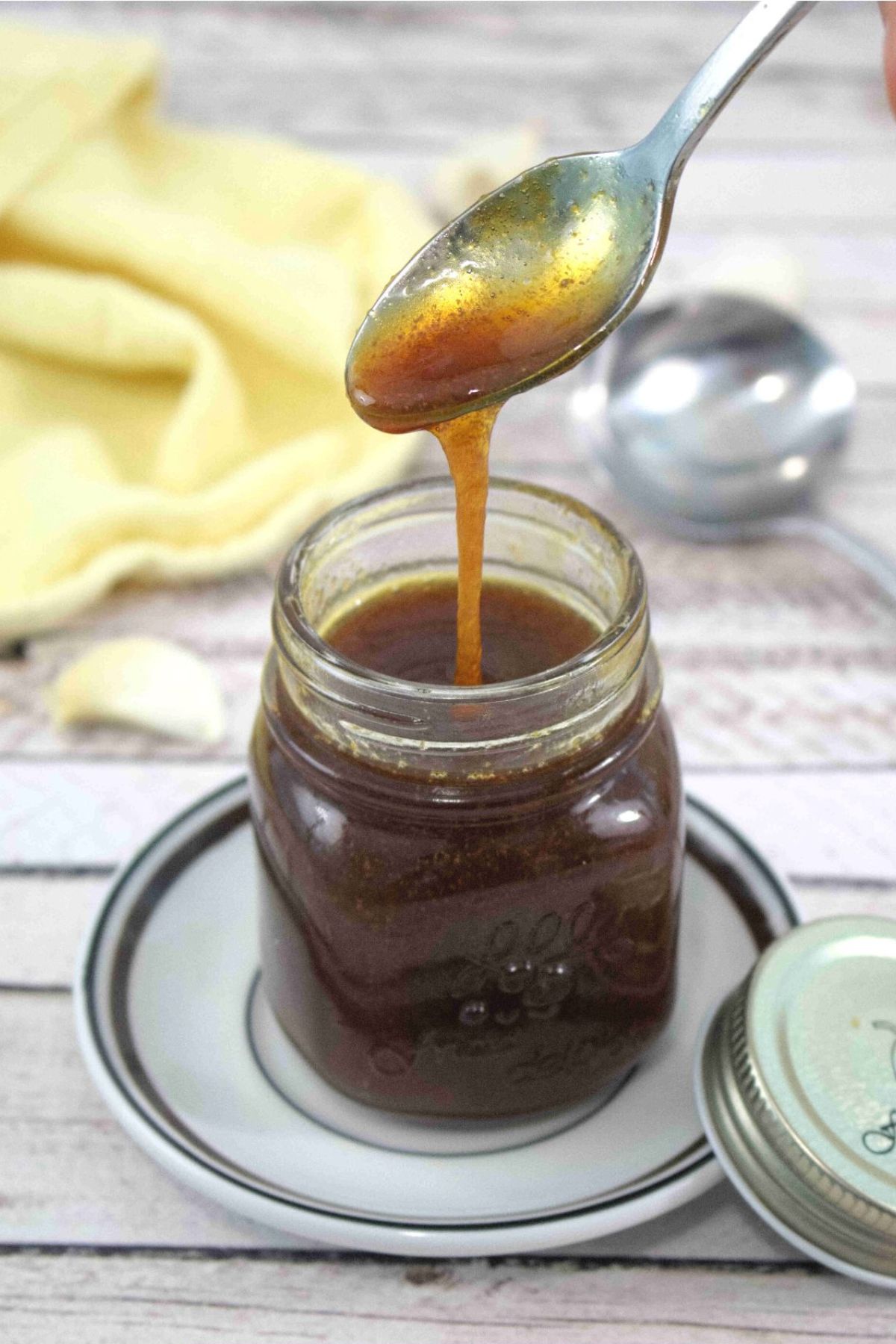 Gluten-free teriyaki sauce in a small jar on a white plate with garlic and a yellow linen around it.