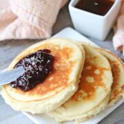 Three yogurt pancakes on a white plate topped with jam. Syrup in a white container behind it and a light orange napkin