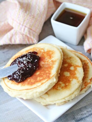 Three yogurt pancakes on a white plate topped with jam. Syrup in a white container behind it and a light orange napkin