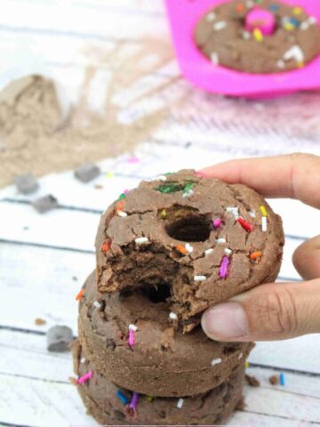 Two fingers placing a chocolate protein donut with sprinkles on top of two more donuts with more behind them.
