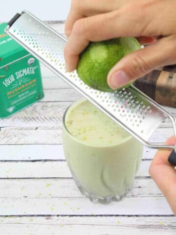 Lime zest being grated over a glass with a green smoothie in it.