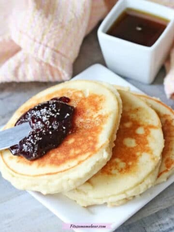Three yogurt pancakes on a white plate topped with jam. Syrup in a white container behind it and a light orange napkin