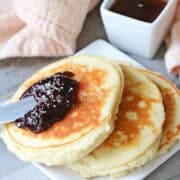 Three yogurt pancakes on a white plate topped with jam. Syrup in a white container behind it and a light orange napkin