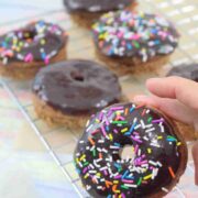 Two fingers holding up a chocolate glazed protein donut with sprinkles with more donuts behind it.