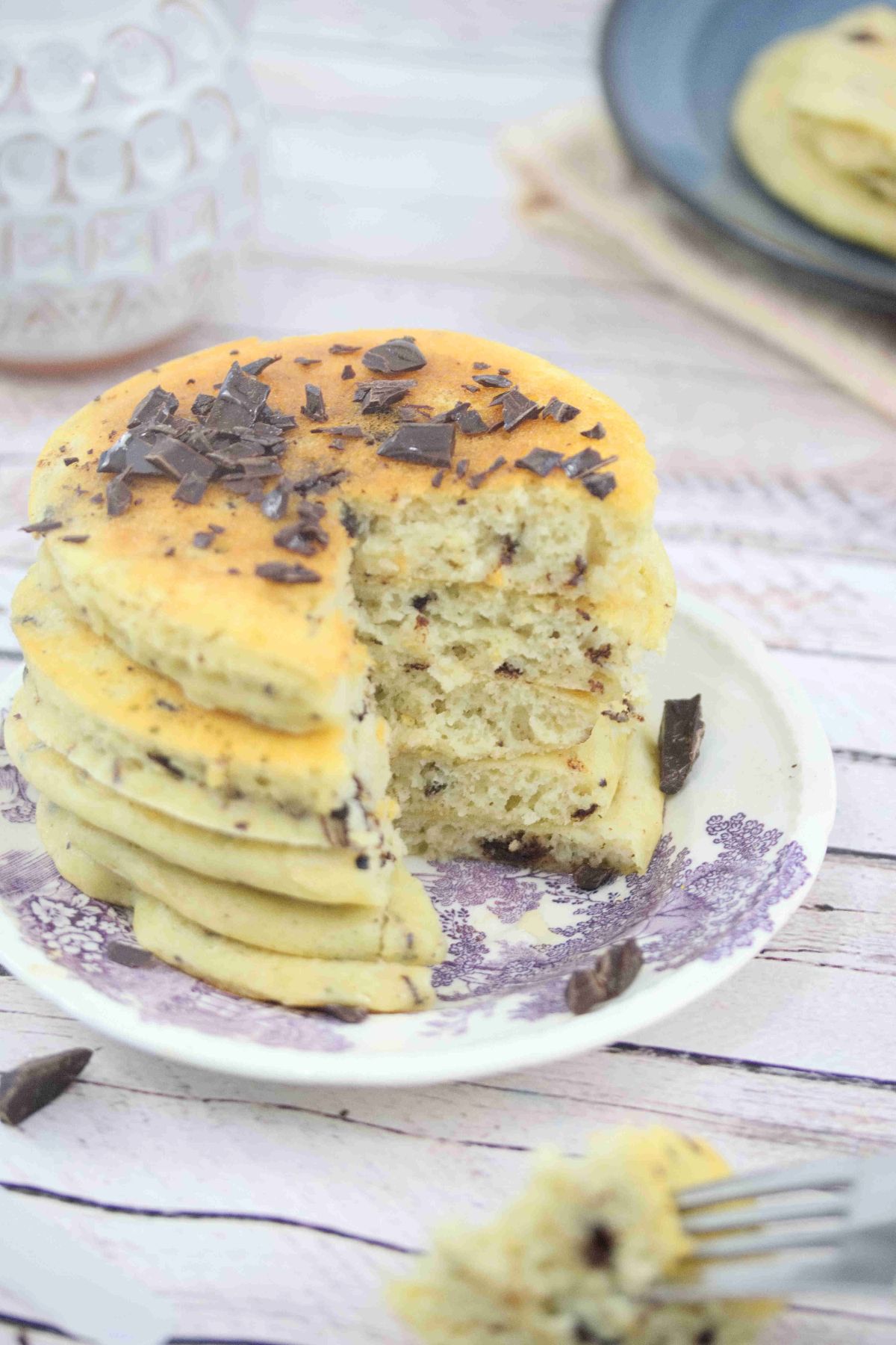 Protein pancakes with chocolate chips stacked on a white and purple platye with a slice cut out of the stack.