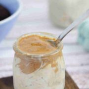 Mason jar with espresso overnight oats topped with almond butter and a spoon in it on a wooden coaster with another jar of oats and a bowl of coffee behind it.