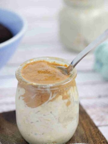 Mason jar with espresso overnight oats topped with almond butter and a spoon in it on a wooden coaster with another jar of oats and a bowl of coffee behind it.