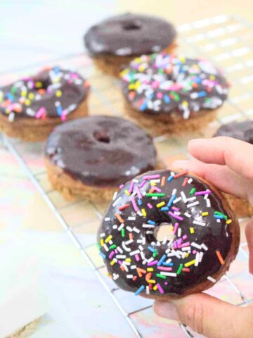 Two fingers holding up a chocolate glazed protein donut with sprinkles with more donuts behind it.