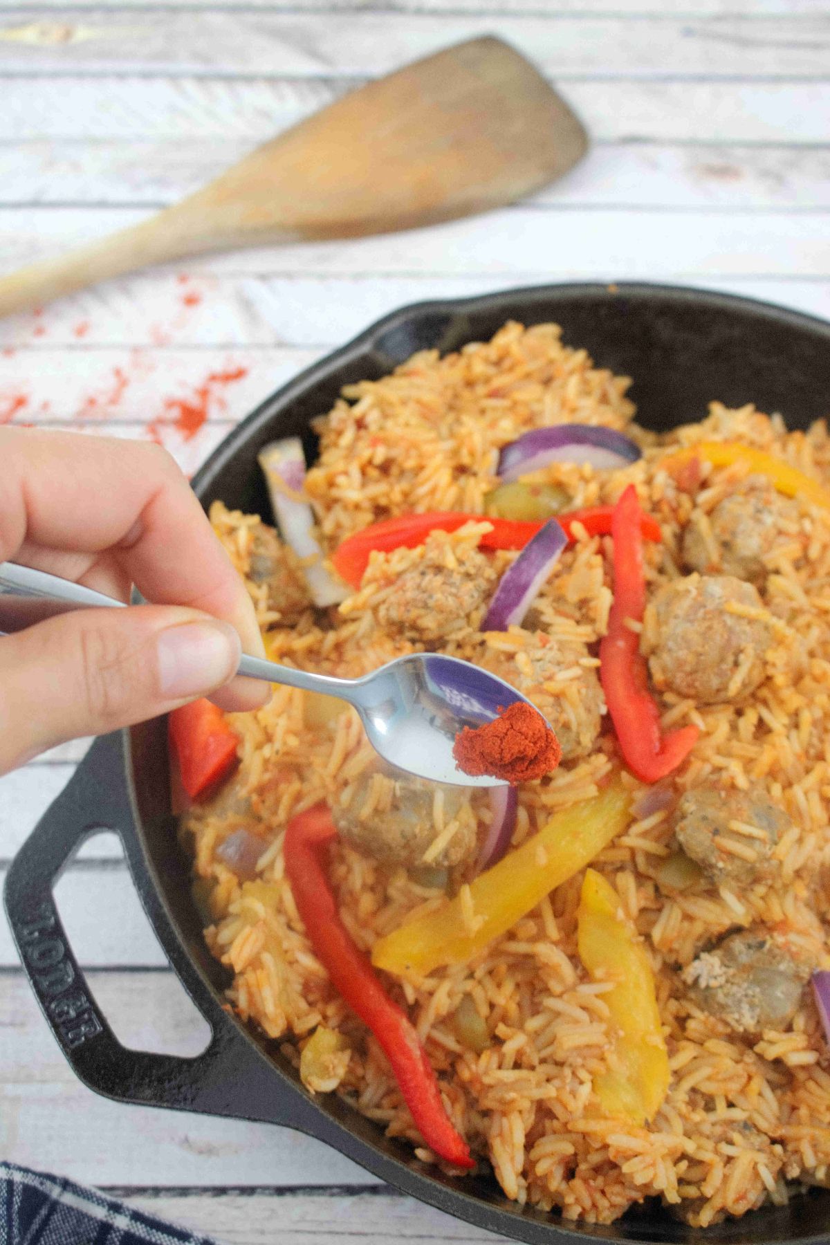 Black skillet with rice, sausages, and peppers in a tomato sauce. and a spoon of paprika being added.