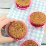 Two fingers holding a protein muffin with a red muffin liner and more banana protein muffins on a blue linen around it.