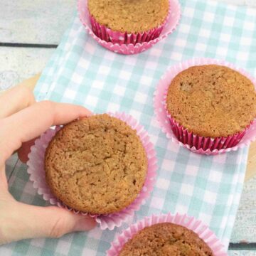 Two fingers holding a protein muffin with a red muffin liner and more banana protein muffins on a blue linen around it.
