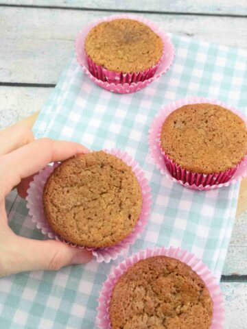 Two fingers holding a protein muffin with a red muffin liner and more banana protein muffins on a blue linen around it.