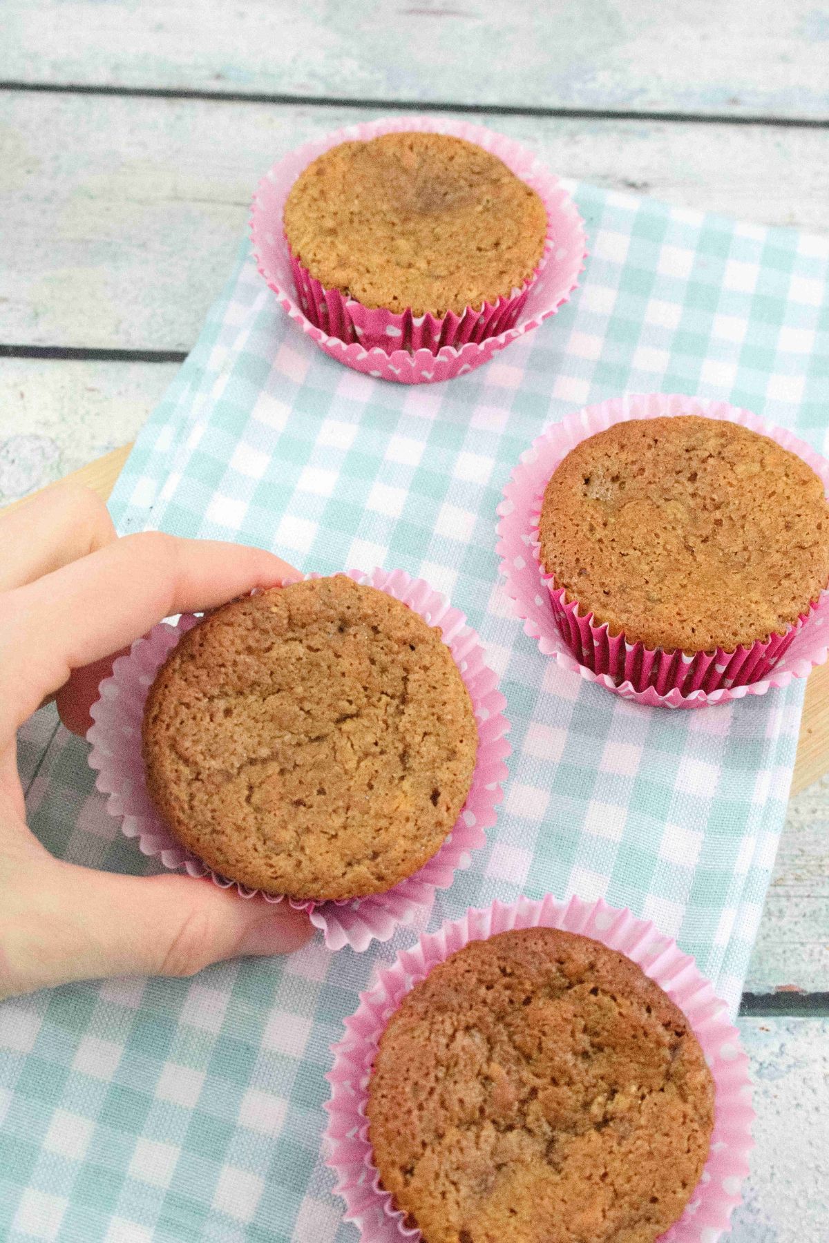Two fingers holding a protein muffin with a red muffin liner and more banana protein muffins on a blue linen around it.