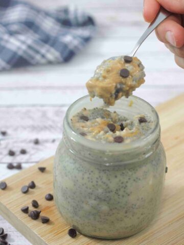 Peanut butter chia pudding with chocolate chips in a mason jar on a cutting board with more chocolate chips around it and a spoon coming out of the mason jar.
